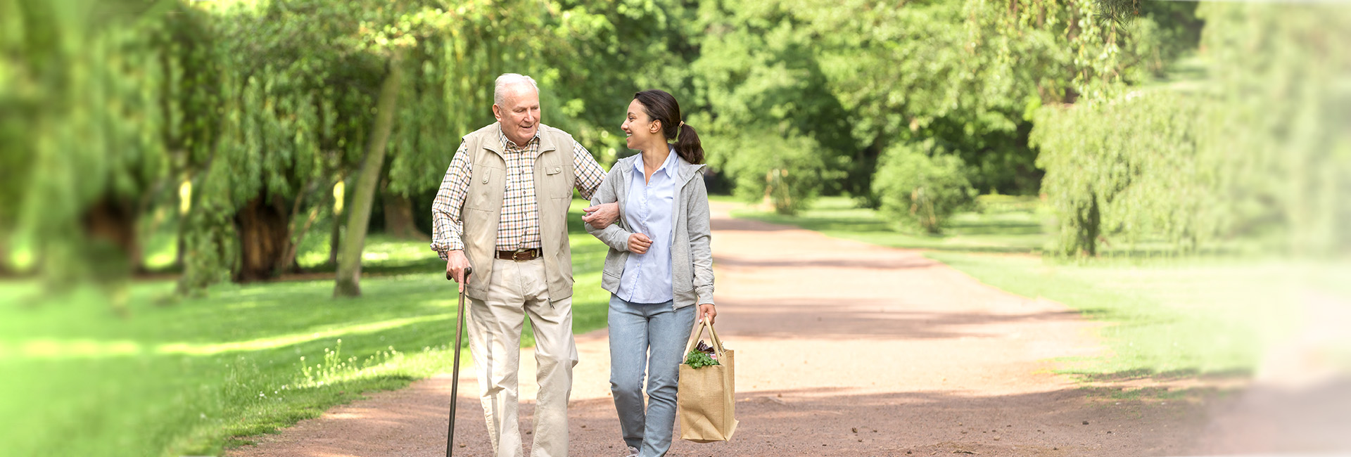 Pflegegeld: Junge Pflegerin spaziert im Park mit altem Mann der am Stock geht