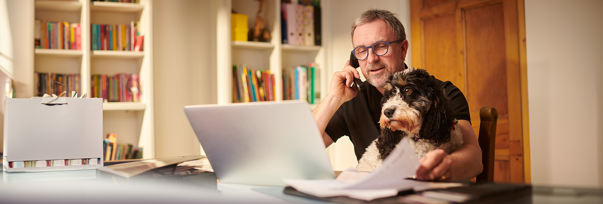 Pensionsantrag: Älterer Mann mit Hund sitzt vor Laptop und telefoniert
