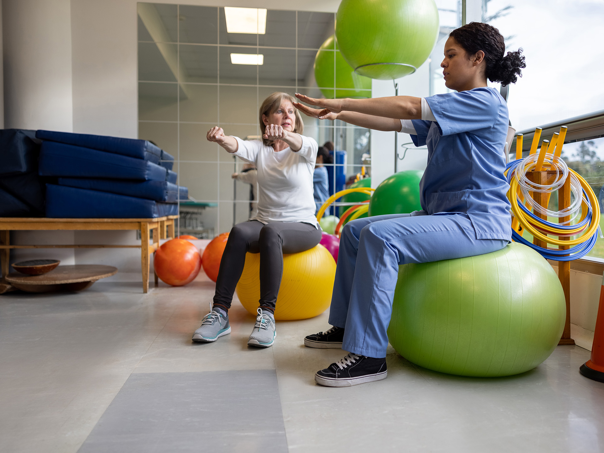 Reha und Beruf: Physiotherapeutin und Patientin in Therapieraum sitzen auf großen Übungsbällen und machen Armübung
