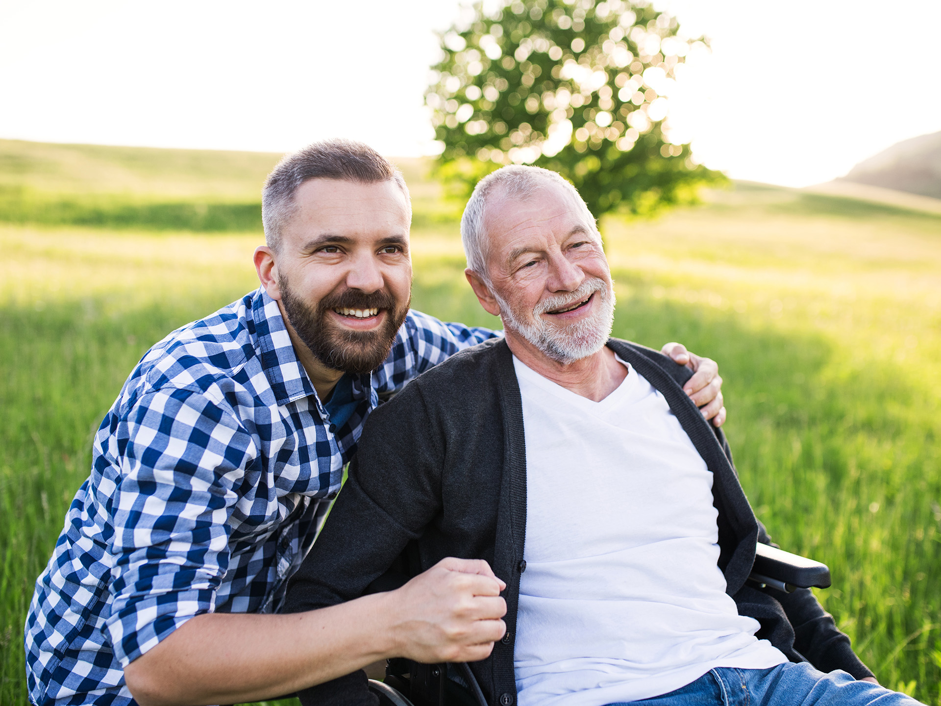 Reha für pflegende Angehörige: Jüngerer Mann schiebt älteren Mann im Rollstuhl vor grüner Wiese